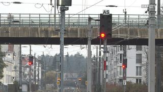 A railway station with multiple poles and electric cables. The train track is visible, and a red light is flashing on a signal pole. Nearby, there are buildings with glass windows and trees.