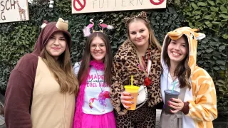 Four women in costumes pose for a photo, with a sign reading 'BITTE NICHT FUTTERN' above them. One wears a giraffe costume, another a bear, one a pink flamingo, and the last a leopard. They all hold drinks.