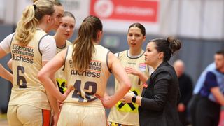 A basketball coach giving instructions to a team of young players in a gymnasium. The team wears yellow jerseys with the word 'BALL' prominently displayed.