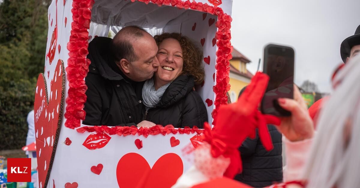 A man and a woman are smiling and kissing inside a heart-shaped booth, surrounded by red hearts and ribbons. They are dressed in warm clothing, and someone is taking a photo of them with a phone.
