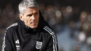 A man with gray hair is wearing a black jacket with white stripes and a white logo on the chest. The background is blurry.