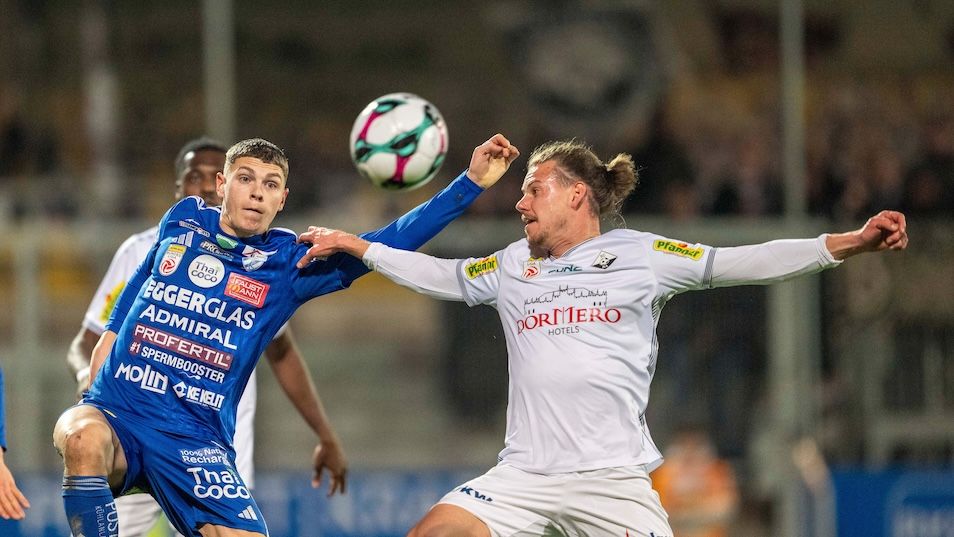 Two soccer players in jerseys clash during a match. One player in blue defends the ball while another in white tries to steal it. The player in blue has visible sponsors' logos.