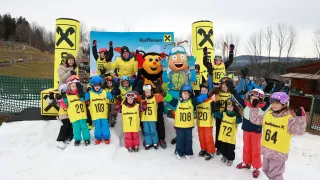 A group of children in numbered ski suits pose for a photo on a snowy day with mascots and adults.