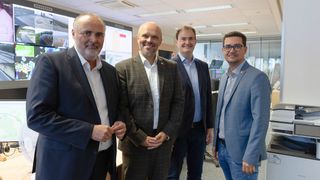 Four men in suits are standing in an office. Three are smiling, one is looking straight ahead. They are surrounded by a large screen and windows.