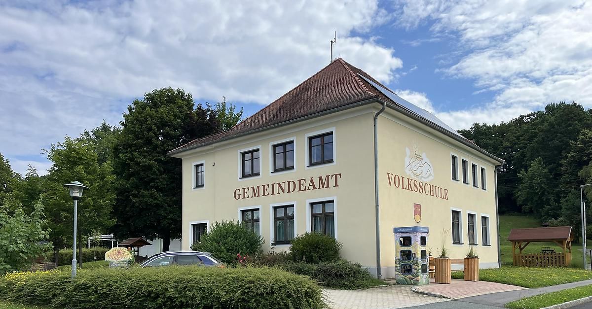 A yellow building with brown roof has German text 'Gemeindeamt' and 'Volksschule'. There are solar panels, windows, and a book exchange box in front. Trees and bushes surround the building. A car is parked on the left.