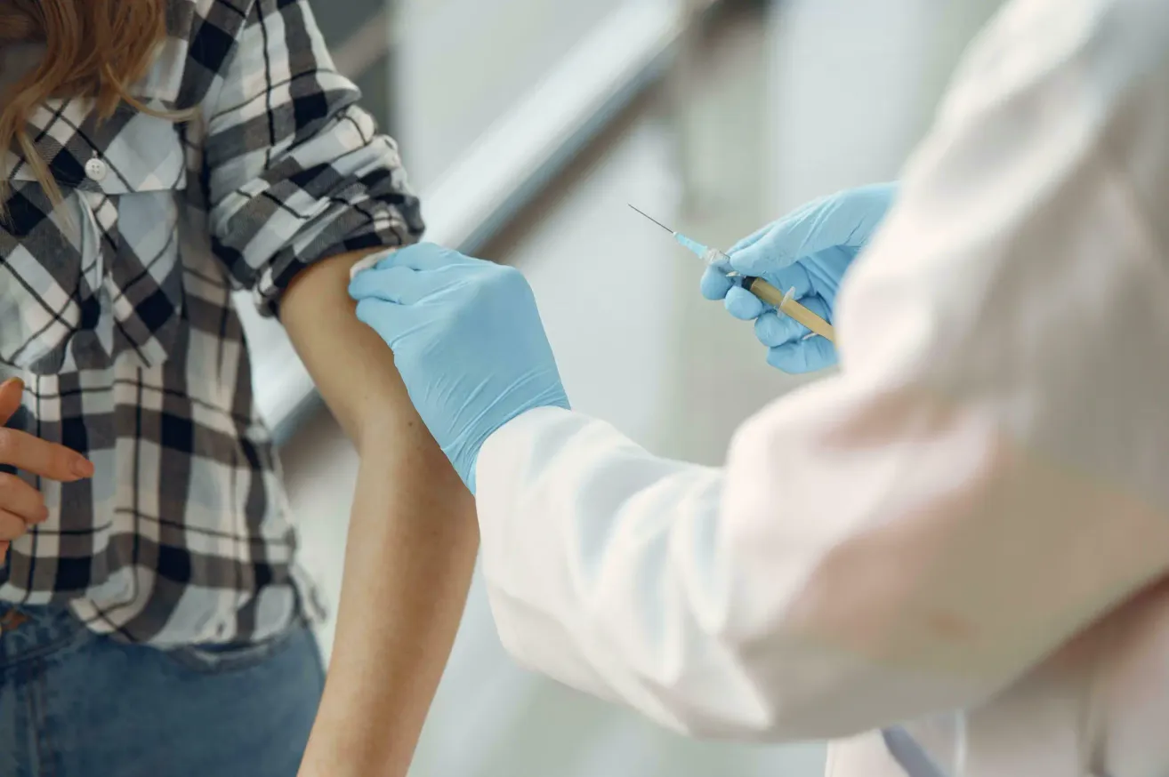 A healthcare worker wearing gloves administers a vaccine to a patient wearing a plaid shirt, using a syringe.