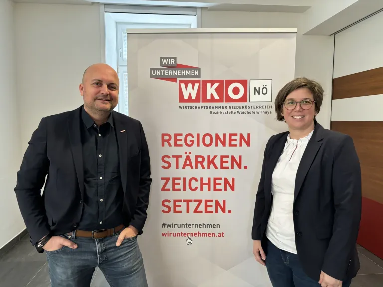 Two individuals stand before a banner, smiling. The banner reads 'Regionen Starken. Zeichen Setzen'. The man wears a black jacket and jeans, and the woman wears a black blazer over a white shirt.