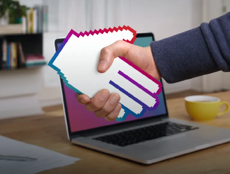 A hand holds a colorful handshake image in front of an open laptop screen on a desk.