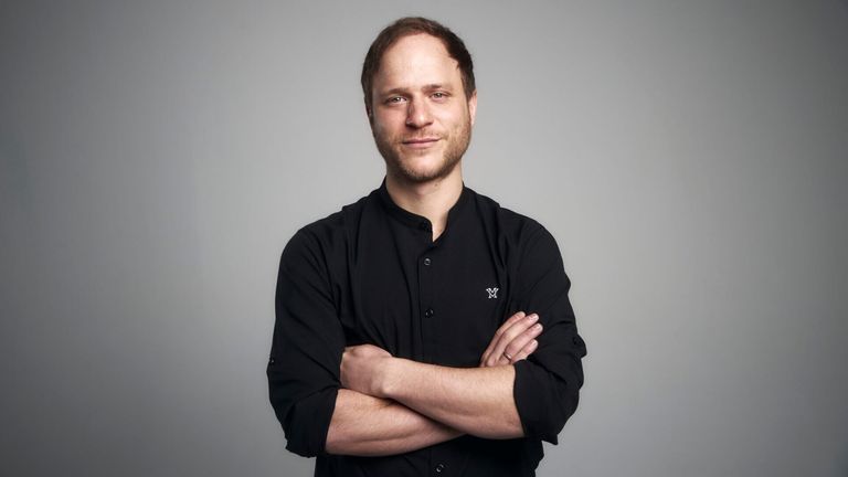 A man with a beard stands with crossed arms in a gray studio. He wears a black shirt with a white logo on the left side of the chest.