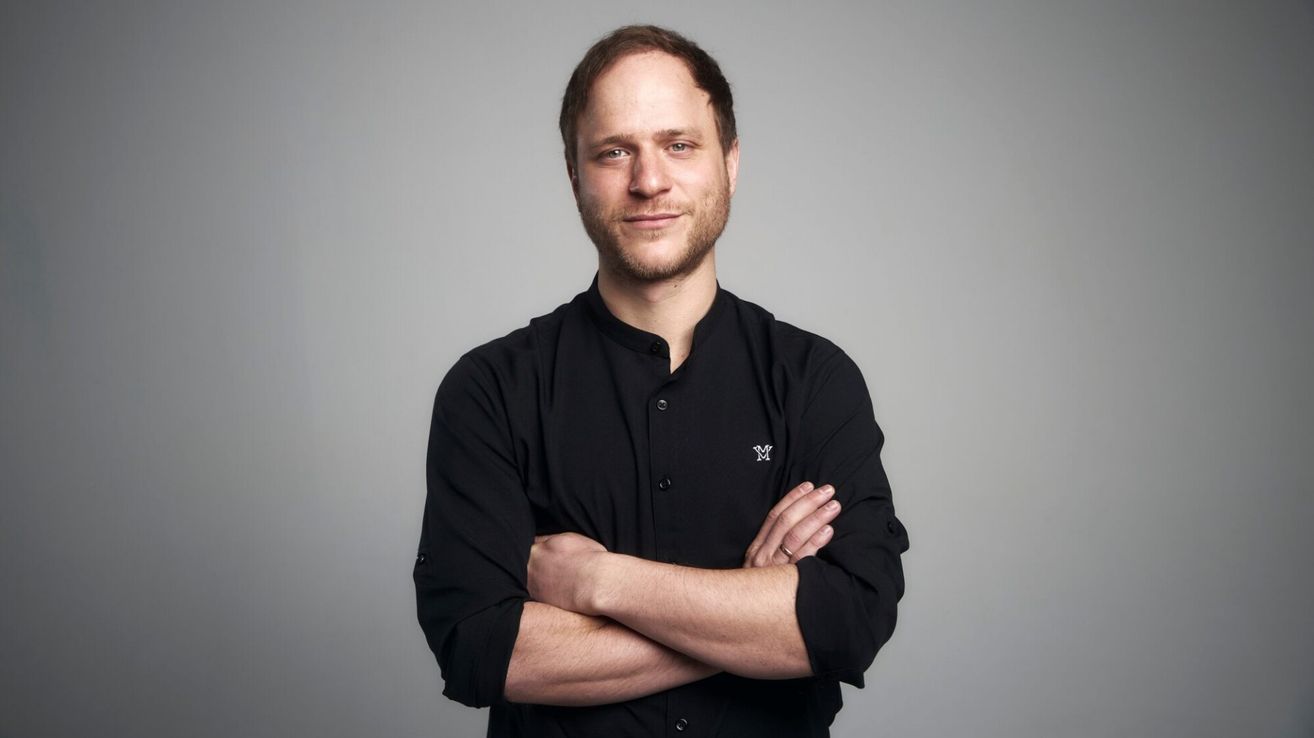 A man with a beard stands with crossed arms in a gray studio. He wears a black shirt with a white logo on the left side of the chest.