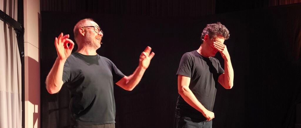 Two men on a stage, one smiling, the other with a hand over his face. Both are wearing black t-shirts and appear to be performing.