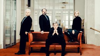 A group of four individuals dressed formally, two men and two women, are seated on a bench in front of a building with glass doors.