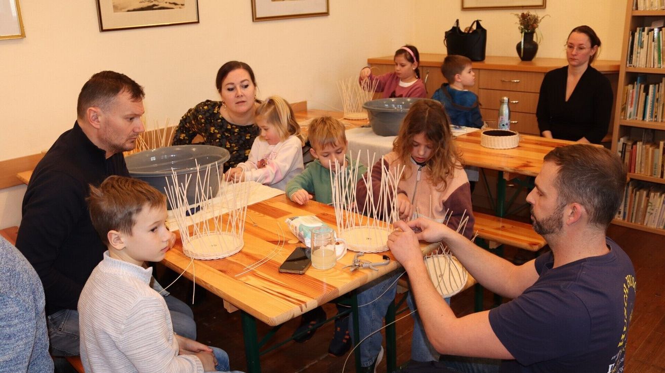 Several children are sitting around a table, crafting with materials like straws and paper. A man and a woman are helping them. The table is cluttered with various crafting items, and there are wooden cabinets and picture frames in the background.