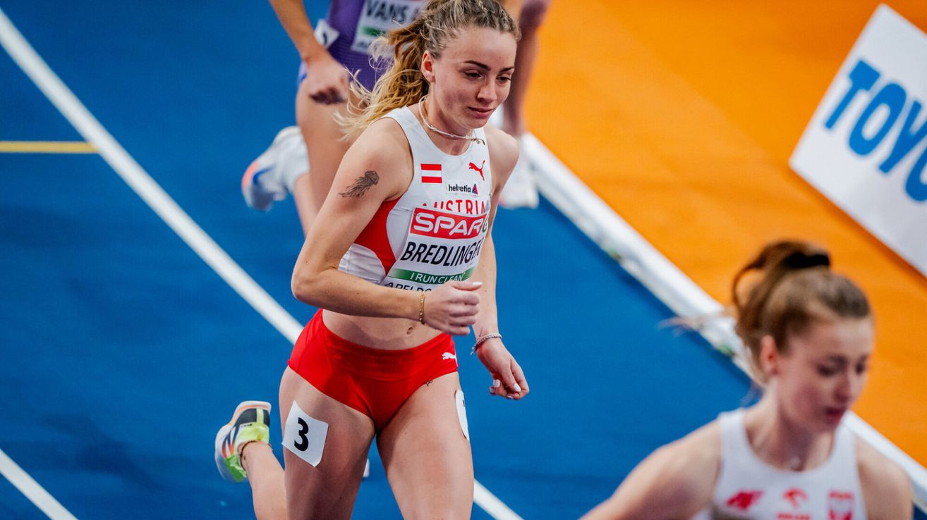 A female athlete with the number 3 on her shorts runs on a track, wearing a white jersey with red accents. She has a tattoo on her arm and is wearing white sneakers. Behind her, another runner is blurry. The background is blue and orange.