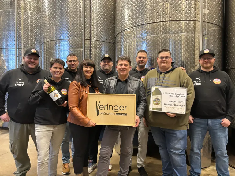 A group of people stands in front of stainless steel barrels. A man holds a plaque and a wine bottle. A woman next to him holds a plaque. They all wear jackets and sneakers. Behind them, a large barrel is visible.
