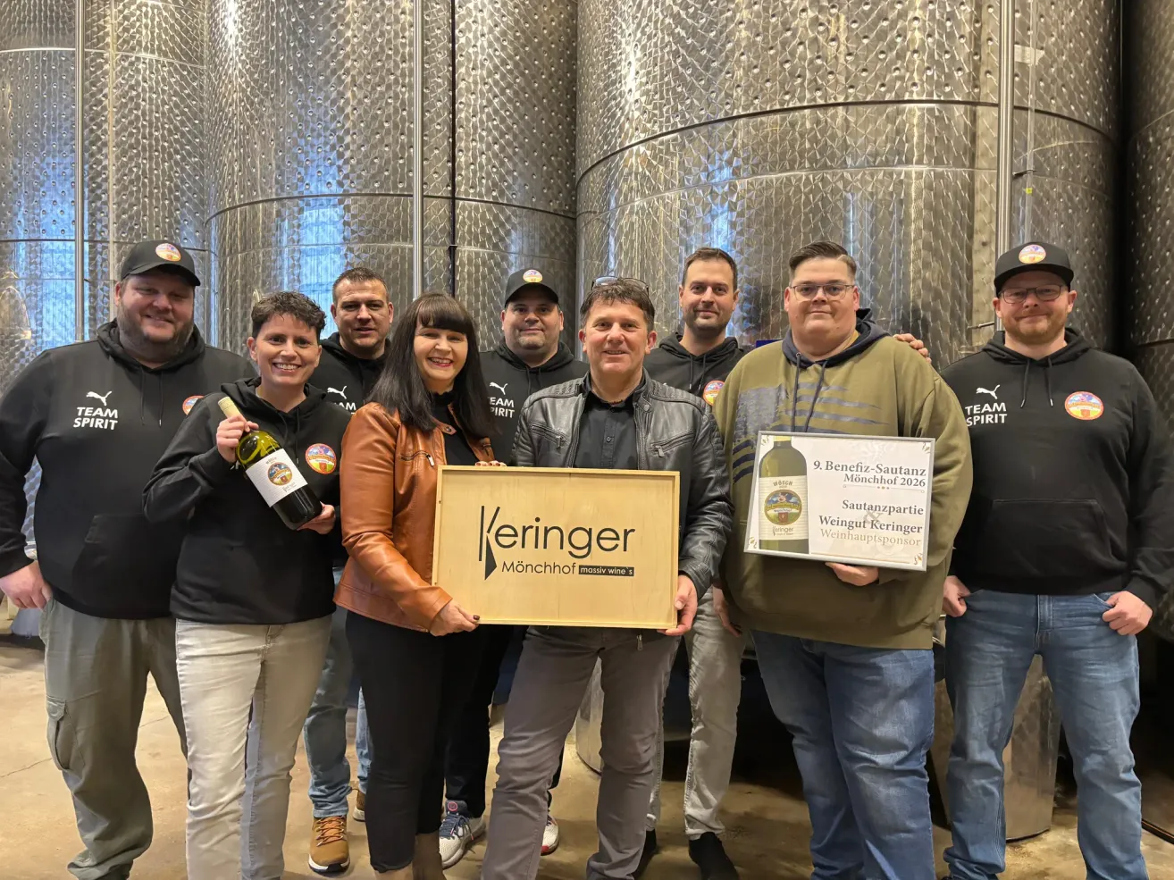 A group of people stands in front of stainless steel barrels. A man holds a plaque and a wine bottle. A woman next to him holds a plaque. They all wear jackets and sneakers. Behind them, a large barrel is visible.