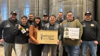 A group of people stands in front of stainless steel barrels. A man holds a plaque and a wine bottle. A woman next to him holds a plaque. They all wear jackets and sneakers. Behind them, a large barrel is visible.
