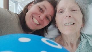Two women are in a hospital bed, smiling and posing for a photo. The woman on the left is younger with dark hair, and the woman on the right is older with white hair.