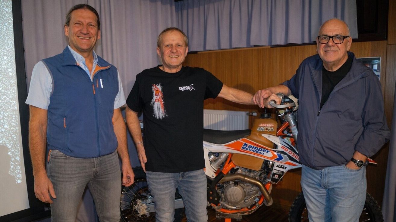 Three men are standing in front of a motorcycle, smiling. The man in the middle is wearing a black shirt with a printed design. The man on the right is holding the handlebar of the motorcycle. The motorcycle has a logo that reads 'Kamp Metalbau' and a number plate that reads '800'.