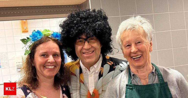 Drei Personen, darunter eine Frau mit blauen und grünen Blumen in den Haaren, ein Mann mit einer Perücke und eine ältere Frau mit einem grünen Schürze, lächeln und posieren für ein Foto.