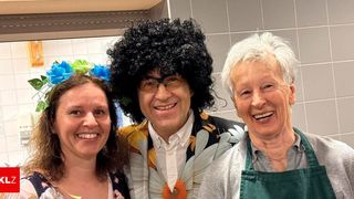 Three individuals, including a woman with blue and green flowers in her hair, a man with a wig, and an elderly woman with a green apron, are smiling and posing for a photo.