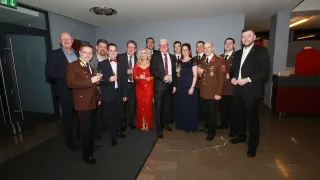 A group of formally dressed people stand in a room, holding wine glasses. A woman in a red dress and a man in a military uniform are among them. They appear to be celebrating an event.