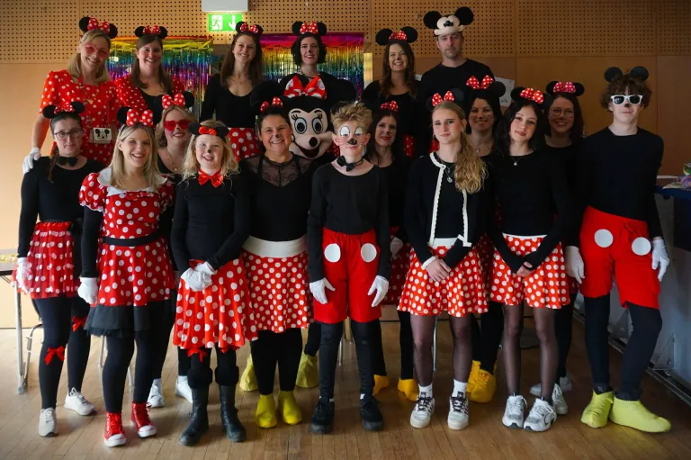 Eine Gruppe von Erwachsenen, die als Minnie und Mickey Mouse verkleidet sind, posiert für ein Foto in einem Raum mit einem mehrfarbigen Hintergrund.