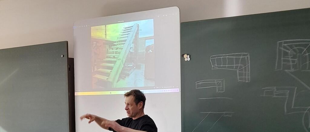 A man is standing in front of a projection screen, showing an image of stairs. He points towards the screen while a blackboard with drawings is behind him.