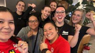 A group of women are smiling and posing for a photo, wearing various colored shirts and glasses. One woman has a necklace and a watch.
