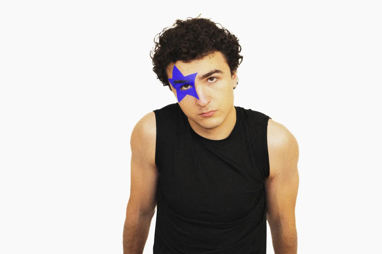 A man with curly hair wearing a black sleeveless shirt has a blue star painted on his face. He is looking to the right with his eyes closed.