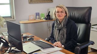 A woman in glasses sits at a desk in an office. She wears a scarf and a watch. The desk has a keyboard, monitor, mouse, and papers. Behind her is a cabinet with books and a framed picture.