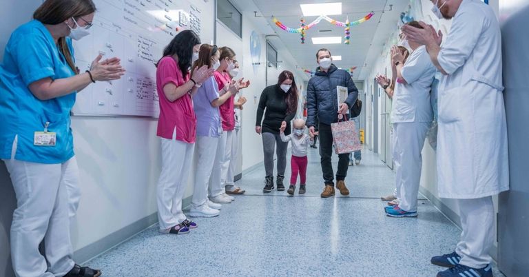 Eine Gruppe medizinischer Mitarbeiter in rosa und weißen Uniformen klatscht, während ein Mann und ein Kind mit einem Beutel durch einen hell erleuchteten Krankenhauskorridor gehen.