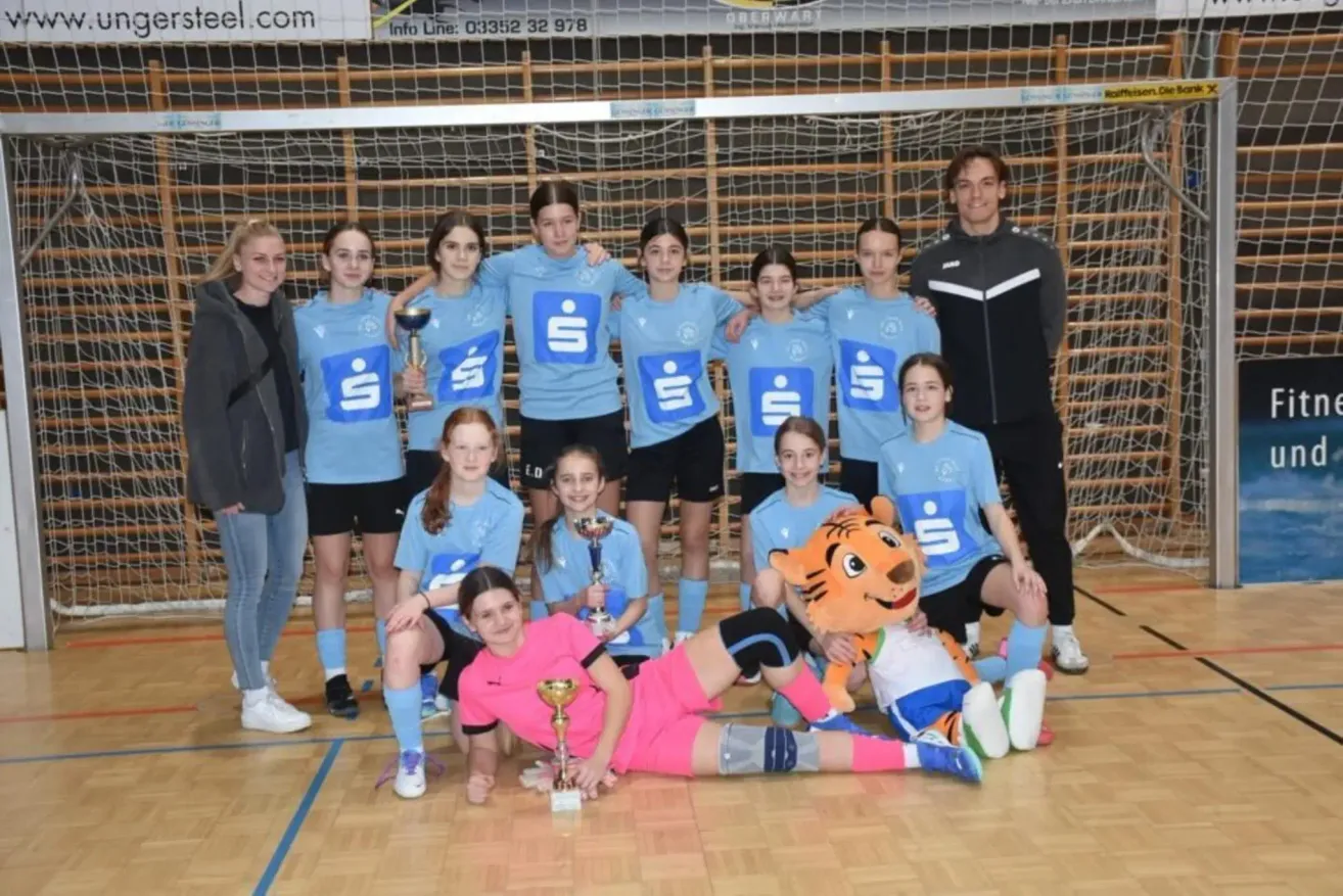 A girls' sports team in blue jerseys poses with a mascot and trophies in front of a goal net.