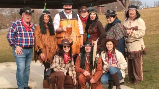 A group of people dressed in various costumes, some holding a glass of beer, pose for a photo in an outdoor setting with a shed in the background.