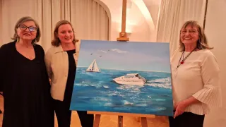 Three women stand next to a painting of a sailboat on the ocean. The painting is on an easel in a room with wooden floors and white curtains.