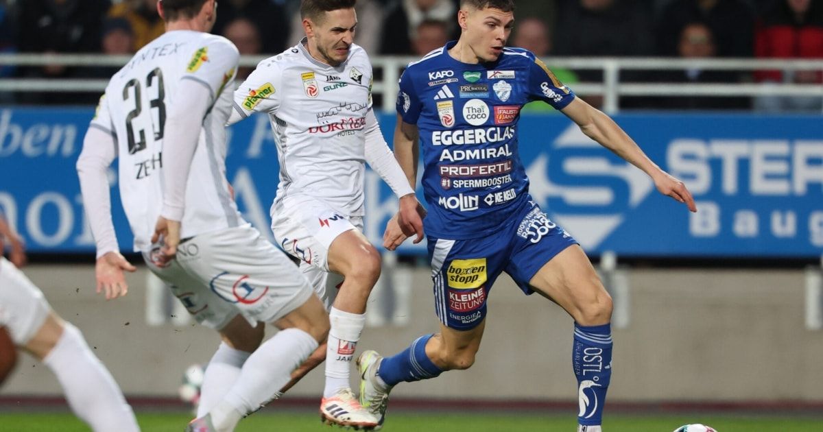 Two soccer players are competing on the field. One is wearing a white jersey, while the other is wearing a blue jersey. Both are sprinting towards the ball.