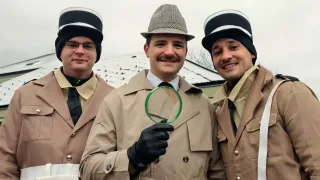 Three men in trench coats and hats pose for a photo, one holding a magnifying glass. They have a cheerful expression.