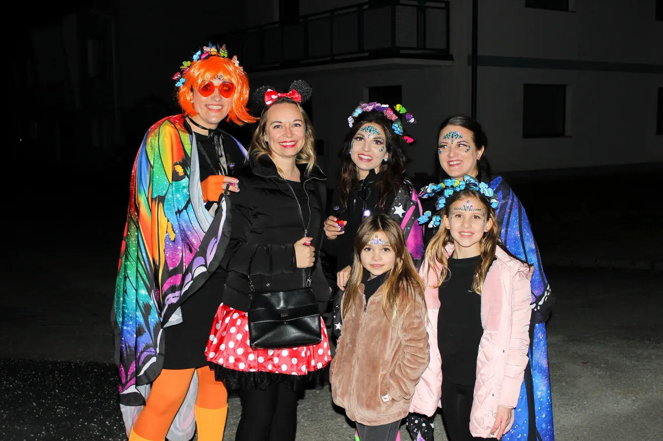 A group of people dressed in colorful costumes and face paint are posing for a photo at night. A woman in a butterfly costume stands beside a girl in a black shirt. Another girl with face paint is holding a cup. Behind them is a building with a balcony.