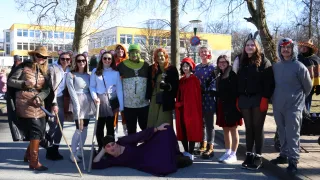 A group of people in costumes are posing for a picture in front of a yellow building. Some wear Mickey Mouse ears, while others have more elaborate costumes.