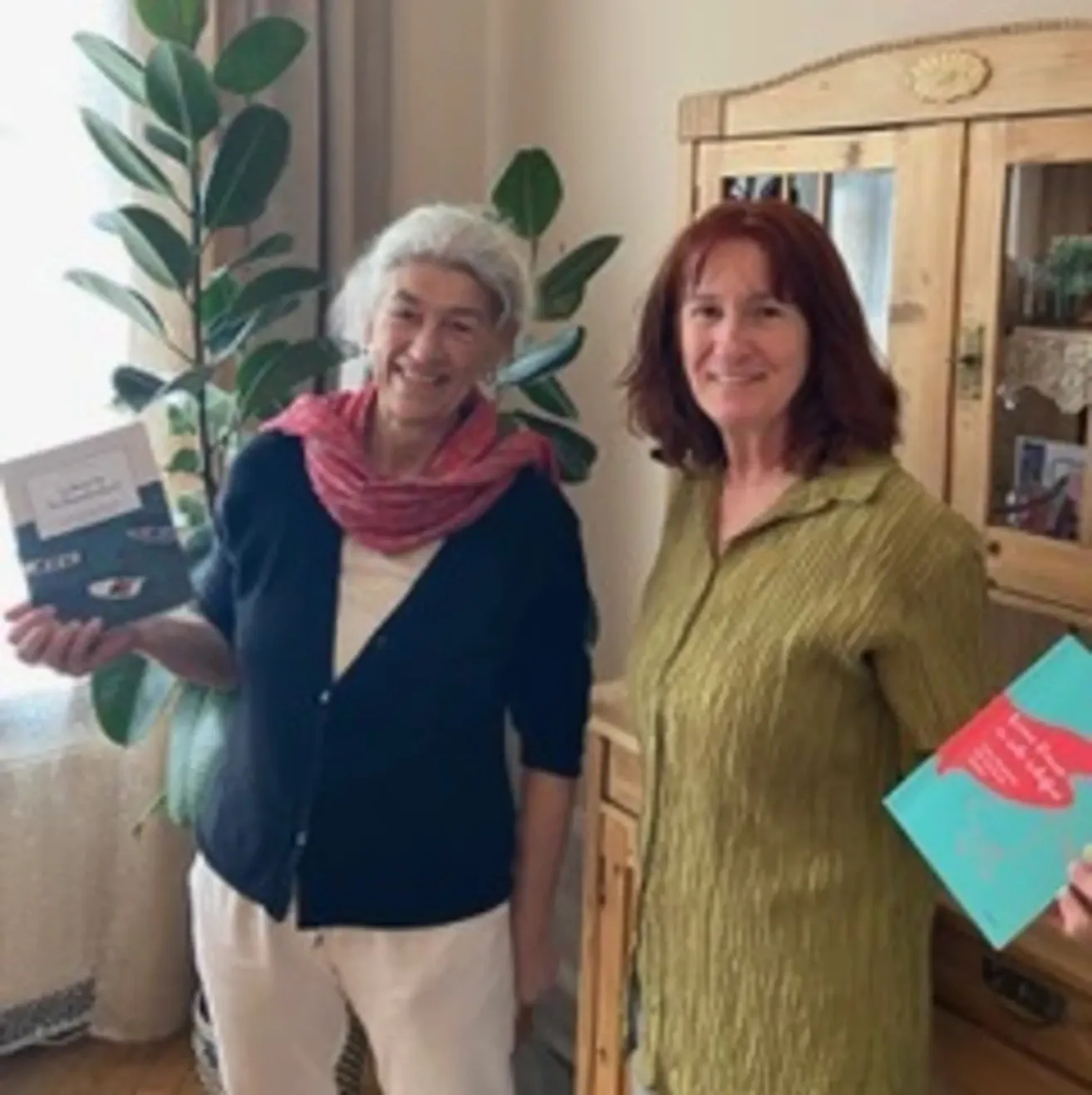 Two women stand together in a room with a cabinet and plant in the background. They hold books and smile at the camera.