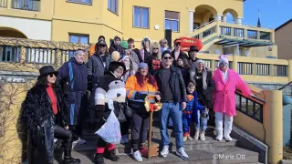 A group of people dressed in various costumes, including a woman in a panda costume, a man in a Superman costume, and a woman in a pink robe, stand on the steps of a building. Behind them is a building with yellow walls and windows.