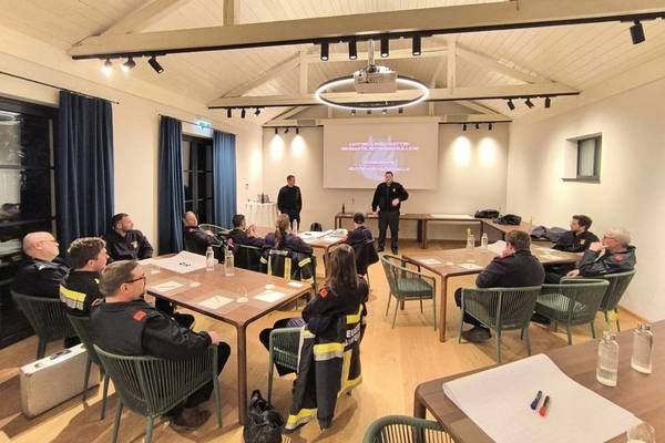 A group of firefighters in uniform are attending a meeting in a room with a projector screen, tables, and chairs. Two people are standing in front of the screen.