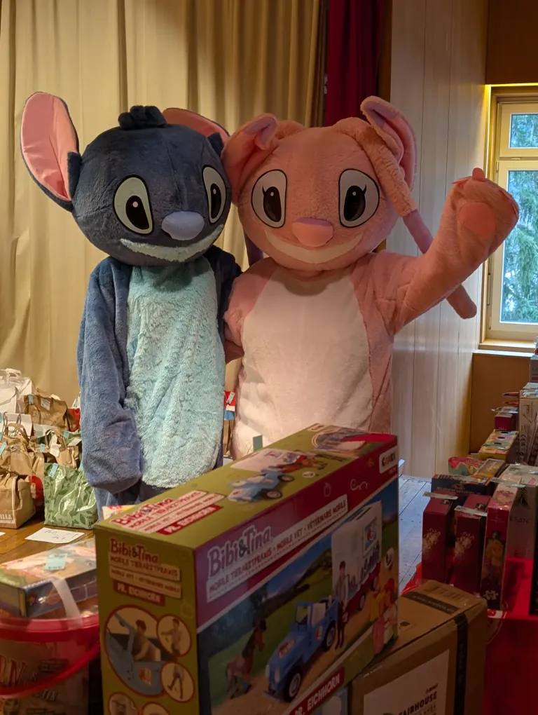 Zwei Maskottchen, eines als Stitch und das andere als rosa Hase gekleidet, stehen neben einem Stapel Geschenkboxen. Die Maskottchen befinden sich in einem Raum mit einer Holzwand und einem Fenster, das Bäume draußen zeigt.