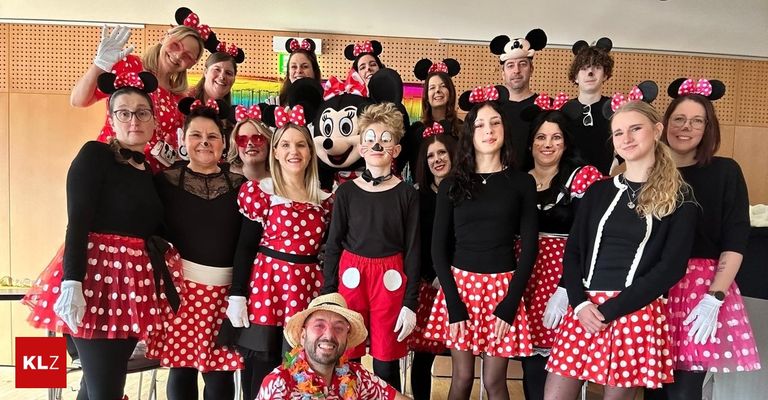 Eine Gruppe von Erwachsenen und Kindern, gekleidet in Micky- und Minnie-Maus-Kostümen, posiert für ein Foto in einem Raum mit einem Micky-Maus-Plüschtier im Hintergrund.