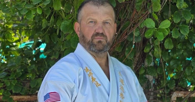 Ein Mann in einem hellblauen Kampfsportanzug mit einem amerikanischen Flaggenaufnäher auf dem Ärmel steht draußen in der Nähe von grünem Laub.