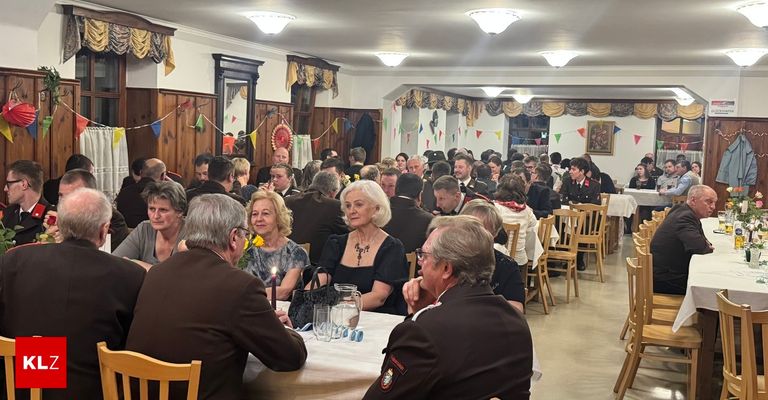 Eine Gruppe von Menschen in formeller Kleidung sitzt an Tischen in einem Bankettsaal, der mit Wimpelketten und gerahmten Kunstwerken dekoriert ist. Eine Frau mit einer gelben Blume hält eine Kerze.