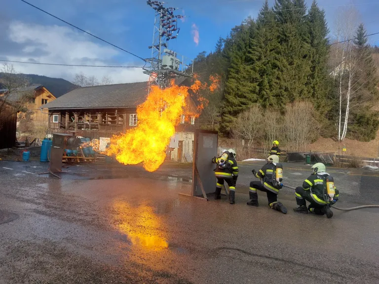 Feuerwehrleute üben das Löschen eines Feuers vor einem Gebäude mit einem Kraftwerk im Hintergrund.
