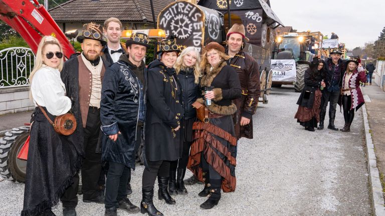 Eine Gruppe von Menschen in Steampunk-Kostümen posiert vor einem Wagen mit der Aufschrift 'Zeitreise Ende' und einem Gebäude im Hintergrund.