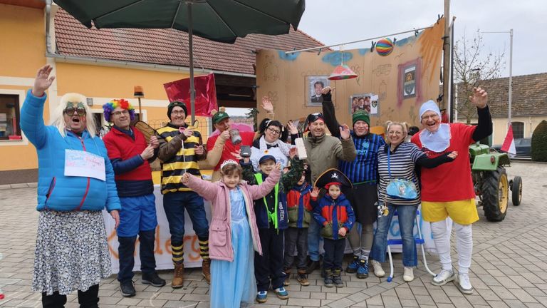 Eine Gruppe von Menschen in Kostümen steht vor einem dekorierten Haus und posiert für ein Foto. Einige tragen Hüte, Brillen und Schuhe. Ein Kind in einem Piratenhut winkt.