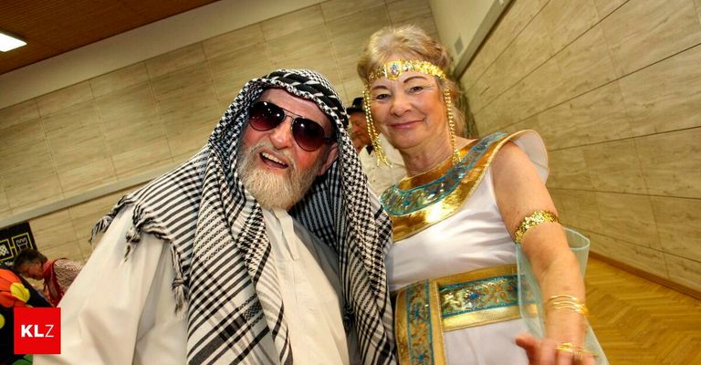 Ein älterer Mann in einem Keffiyeh und Sonnenbrille steht neben einer Frau in einem altägyptischen Kostüm, beide lächeln für ein Foto in einer Halle.